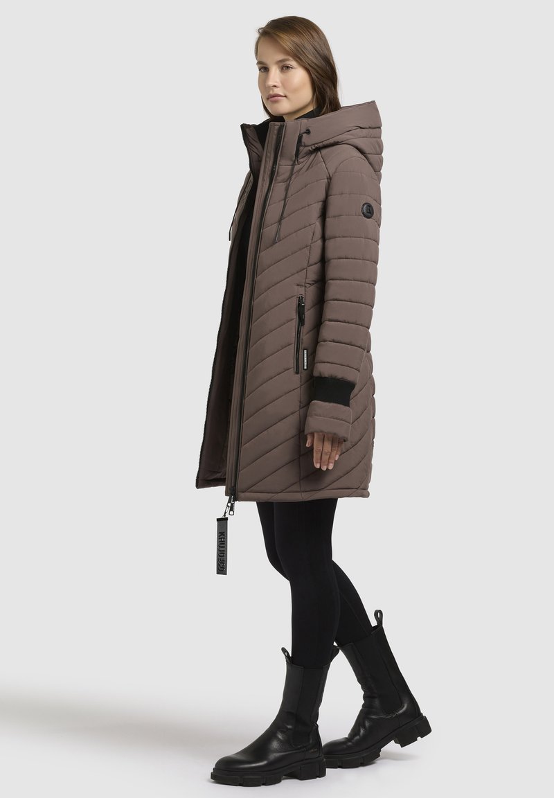 Abrigo acolchado marrón con capucha, con detalles en negro, bolsillos frontales con cremallera y puños acanalados. Llevado sobre leggings negros y botas.