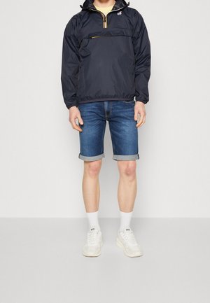 Marinblå vattentät pulloverjacka med halvlång dragkedja, gula detaljer och framficka, kombinerad med denimshorts och vita sneakers.