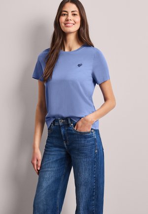 Mujer de pie con una mano en el bolsillo, vistiendo una camiseta de manga corta azul claro con logo de corazón y vaqueros de mezclilla azules sobre un fondo liso.