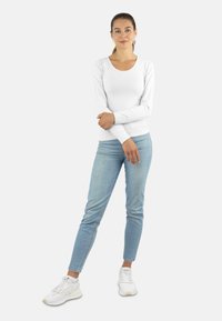 Langarm weißes Oberteil mit rundem Ausschnitt, kombiniert mit hellblauen Skinny-Jeans und weißen Sportschuhen, vor einem schlichten Hintergrund stehend.