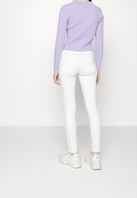 Lila ribbad långärmad topp som kombineras med vita tajta jeans och kraftiga vita sneakers. Bakgrunden är helt vit, vilket fokuserar på outfiten.