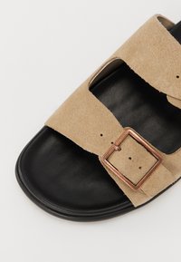 Beige semsket sandal med to brede stropper, utskjæringer, og en metallisk kobberbeltespenne på en svart lærsåle. Glatt tekstur, minimalistisk design.
