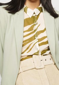 Ljusgrön kavaj över en krämfärgad skjorta med olivgröna zebraränder, kombinerad med beige manchesterbyxor och ett matchande bälte.