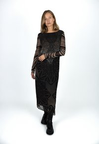 Langes schwarzes Kleid mit transparenten langen Ärmeln, verziert mit floralen und geometrischen Mustern, getragen mit schwarzen klobigen Stiefeln. Glatte Textur, elegantes Design.