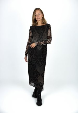 Long black dress with sheer long sleeves, featuring floral and geometric patterns, worn with black chunky boots. Smooth texture, elegant design.
