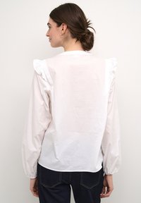 Blusa bianca a maniche lunghe con dettagli arricciati sulle spalle, vestibilità comoda e tessuto morbido e leggero; abbinata a pantaloni in denim scuro.