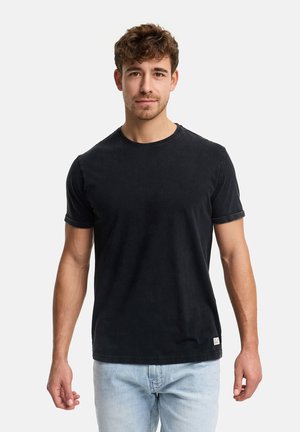 Junger Mann mit kurzem lockigem Haar, der ein schwarzes T-Shirt mit kurzen Ärmeln und hellblaue Jeans trägt, vor einem schlichten weißen Hintergrund stehend.
