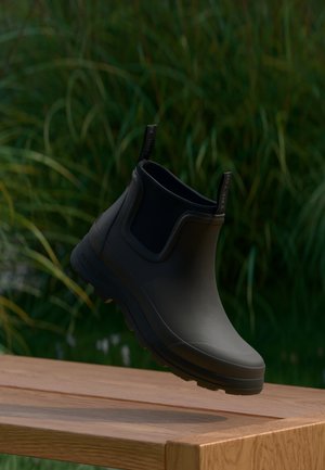 Zwarte rubberen enkellaars met een gladde afwerking en een getextureerde zool, voorzien van een trekkoord voor gemakkelijk aantrekken, weergegeven tegen een vervaagde groene achtergrond.