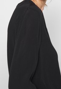 Maglietta nera a maniche lunghe in un tessuto morbido, con una vestibilità comoda e un design minimalista, caratterizzata da una silhouette leggermente drappeggiata e senza decorazioni visibili.
