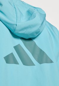 Une veste bleu clair avec une texture lisse présente un grand design de logo Adidas bleu foncé au dos, avec un détail de capuche.