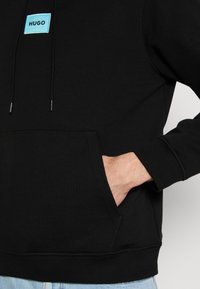 Sudadera negra con una etiqueta turquesa que muestra "HUGO." Incluye un bolsillo frontal, puños acanalados y una tela suave y texturizada.