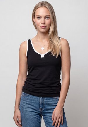 Junge Frau mit langen blonden Haaren, die ein schwarzes Tanktop mit weißem Besatz und blaue Jeans trägt, steht vor einem schlichten hellen Hintergrund.