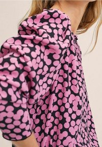 Løs pasform bluse i marineblå med et pink prikmønster. Har pufærmer og en samlet halsudskæring. Glat, let stof.