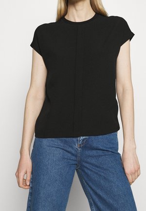 Femme portant un haut noir uni à manches courtes et un jean bleu taille haute sur fond blanc.