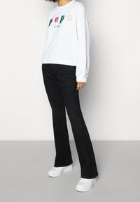 Persona vestida con sudadera blanca con el logo multicolor "PEPE Jeans", jeans acampanados negros y zapatillas plataforma blancas, de pie sobre un suelo gris.