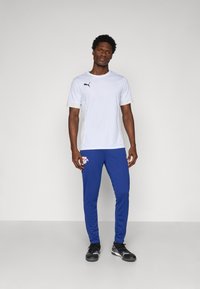 Camică sport albă cu mânecă scurtă, având un logo negru, asociată cu pantaloni albastre ce includ un logo. Materialul pare ușor și respirabil.