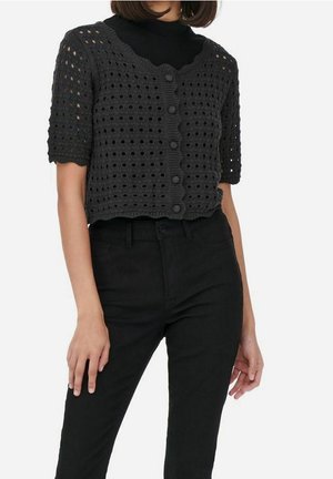 Femme portant un cardigan en crochet noir à manches courtes avec boutons par-dessus un haut noir, associé à un pantalon noir taille haute.