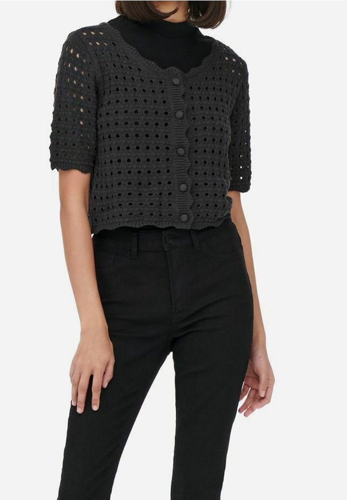 Femme portant un cardigan en crochet noir à manches courtes avec boutons par-dessus un haut noir, associé à un pantalon noir taille haute.