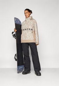 Snowboard z czarną bazą i niebieskim akcentem; model w beżowej kurtce w kwiaty i czarnych spodniach, trzymający deskę, na tle jednolitego tła.