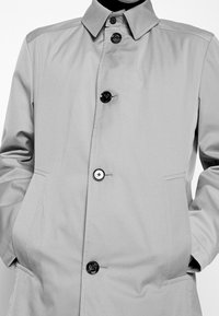 Cappotto lungo grigio chiaro, a maniche lunghe, realizzato in tessuto morbido, con colletto a punta, chiusura con bottoni e due tasche laterali. Bottoni neri e dettagli di cucitura.