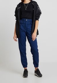 Marinblå denim joggers med elastiska muddar, hög midja, två framfickor, och bärs med en svart topp och jacka.