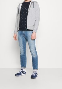 Grauer Zip-Hoodie mit Kordeln, navyfarbiges T-Shirt mit Muster, hellblaue Jeans und navyfarbige Sportschuhe mit weißen Akzenten.