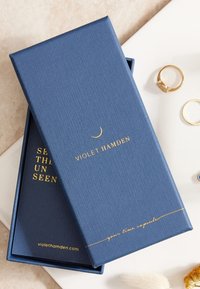 Blaues, strukturiertes Kästchen mit goldgeprägtem Text. Enthält eine schwarze Innenbox mit goldener Schrift. Umgebende goldene Ringe sind sichtbar.