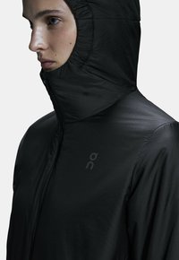 Black lightweight jacket with a high collar and fitted hood. Features a smooth texture and a minimalistic logo on the chest.