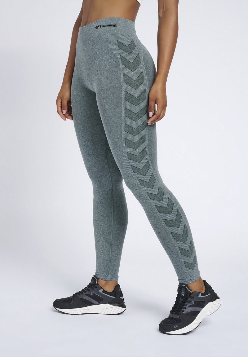 Woman wearing high-waisted grey leggings with dark chevron stripes on sides and black athletic shoes on light background.