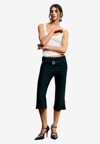 Zwarte capri-broek met een uitlopende zoom en een riem met een gouden D-vormige gesp, gecombineerd met een wit tanktop en zwarte hoge hakken.