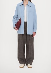 Světle modrá košilová bunda, bílý top, hnědé kostkované široké kalhoty a krémové tenisky. Drží vínově červenou psaníčko.