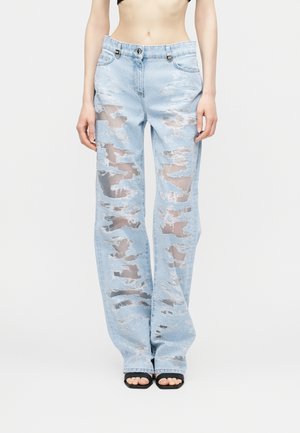 Φως μπλε τζιν με μεγάλες οριζόντιες σχισμές που αποκαλύπτουν το δέρμα από κάτω, φορεμένα με μαύρα σανδάλια ανοιχτής μύτης σε μοντέλο που στέκεται.