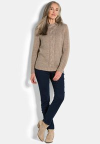 Pull en maille beige avec un motif en torsades, associé à un pantalon bleu foncé et des bottines beiges, se tenant devant un fond uni.