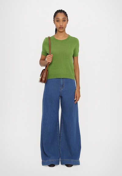 Grøn kortærmet sweater parret med brede blå denimbukser. Modellen holder en brun crossbody taske med en rem. Glat tekstur overalt.