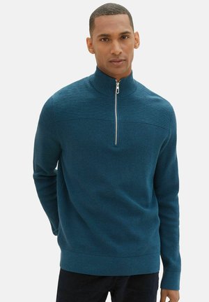 Suéter azul verdoso con cremallera, textura acanalada y detalle de rayas horizontales en el cuello y la parte superior del pecho. Ajustado, de mangas largas y acabado suave.