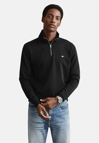 Pullover nero con mezza cerniera, realizzato in tessuto strutturato, con un piccolo logo sul petto. Abbinato a jeans azzurri.