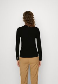Femme aux cheveux bouclés portant un haut noir ajusté à manches longues et un pantalon couleur camel, debout face à un mur blanc uni.