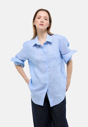 Femme portant une chemise bleu clair à boutons avec les manches retroussées et un pantalon sombre, debout les mains sur les hanches devant un fond uni.