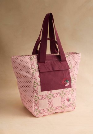 Vaaleanpunainen kangaskassi mansikkakuvioilla, tumman vaaleanpunainen etutasku ja siihen sopivat kahvat neutraalia taustaa vasten.