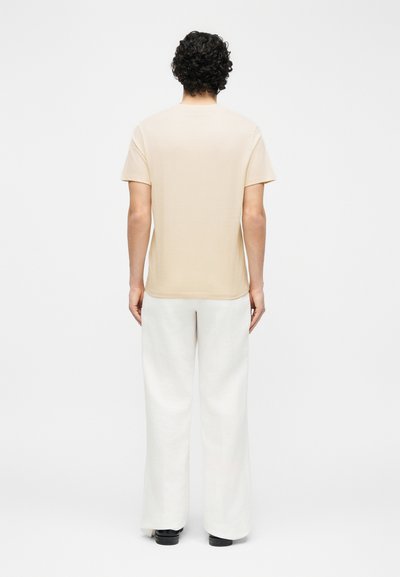 Άτομο με σγουρά μαλλιά που φοράει μπεζ κοντομάνικο πουκάμισο, λευκό παντελόνι φαρδιάς γραμμής και μαύρα παπούτσια, όρθιο και με την πλάτη στραμμένη σε λευκό φόντο.