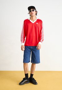 Röd långärmad tröja med vit krage och tre vita ränder på varje ärm, tillsammans med blå denimshorts och svarta sneakers.