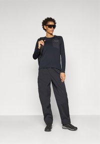 Maglietta nera a maniche lunghe con taschino sul petto, abbinata a pantaloni cargo neri larghi. L'outfit include occhiali da sole neri e scarpe da trekking.