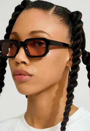 Črna pravokotna sončna očala z oranžno obarvanimi stekli, s krepko oblikovanim okvirjem in sijajnim zaključkom. Lasje so oblikovani v zvite kite.