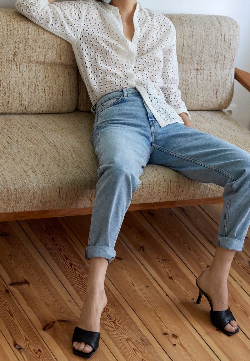 Chemise blanche à œillets, jeans bleu clair à revers, et mules noires à talons ouverts. Canapé avec un tissu beige texturé, cadre en bois.