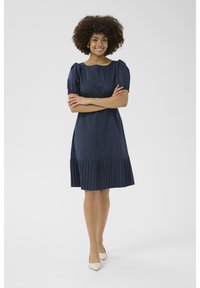 Námornícky modré krátke šaty s puffovými rukávmi a plisé spodným častiam, vyrobené z hladkej tkaniny; model pózuje so skríženými rukami na neutrálnom pozadí.