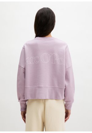 Osoba z długimi ciemnymi włosami, ubrana w liliową bluzę z napisem „Marc O’Polo” na plecach oraz beżowe spodnie, zwrócona tyłem.