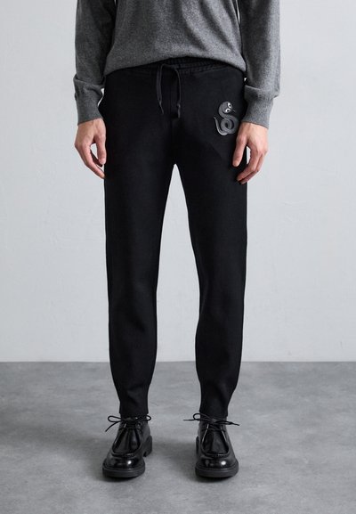 Pantaloni de trening negri cu talie reglabilă și buzunare laterale. Prezintă un design texturat în formă de șarpe pe piciorul stâng. Asortați cu încălțăminte neagră cu șireturi.