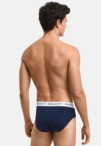Marineblaue Unterhose mit einem weißen Gummibund, der den wiederholten Text "GANT" trägt, und einem glatten, figurbetonten Design.