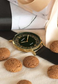 Montre à bracelet en maille noire avec un boîtier doré, cadran vert pailleté, index horaires dorés et le nom de la marque "OOZOO Vintage." Entourée de cookies.