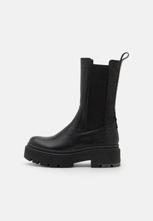 Bottine noire à talon avec une semelle épaisse compensée, présentant une finition en cuir lisse et des panneaux latéraux élastiques pour faciliter l'enfilage.